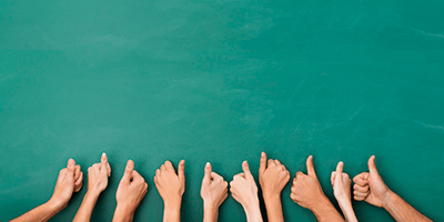 Close-up-view-of-the-hands-of-a-group-of-people-giving-a-thumbs-up-gesture-of-approval-an-success-with-their-hands-raised-against-a-blank-green-chalkboard-with-copyspace